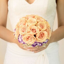 This image offers a detailed, midsection view of a bride holding her bouquet at chest height, as if she has just stepped out from a ceremony at Stanmore Station's nearby church or a hidden garden venue off Marsh Lane. Her wedding gown is a gentle ivory or pure white, with fine lace tracing the bodice and a soft V-neck that feels both traditional and contemporary. The fabric below the waist gathers in gentle folds, the texture suggesting a light, flowing material that moves easily as she walks. In her hands rests a meticulously crafted, round bridal bouquet that draws the eye with its harmony of colour and form. The upper portion forms a plush dome of roses in soft peach, apricot, and blush pink tones, each bloom plump and closely nestled, petals curling inward like layers of silk. These warm shades catch the ambient light, giving the bouquet a luminous, sunrise-like glow. Just beneath this central cluster runs a striking ring of deep purple flowers edged in creamy white ruffles, likely lisianthus, creating a rich contrast that makes the roses appear even softer. Around the base, delicate sprays of tiny white filler flowers-reminiscent of baby's breath-add a misty, romantic haze and soften the transition to the bride's dress. Her nails are neatly manicured in a pale, understated polish, with a subtle hint of glitter on one thumb that adds a personal sparkle, as if ready to catch in a photographer's flash. The photo radiates a gentle, romantic calm, making it perfect for illustrating articles about bridal bouquets, wedding flower trends in Stanmore, or how local florists curate colour palettes that feel intimate yet elegant for North London couples.