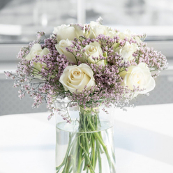 This photograph presents a tender, contemplative bouquet composed of soft white roses and lavender-pink filler flowers, artfully arranged to convey sympathy and calm for recipients around Stanmore. The white roses, fully open and generously petaled, take center stage; their creamy tones and spiral hearts suggest purity, remembrance, and an unspoken gentleness. Each rose appears velvety and full, with petals cupping one another in layers that seem almost protective. Resting delicately between the roses are sprigs of statice, their tiny blossoms in hues of pale lilac, dusky purple, and a touch of blush softening the strong, pure white of the main blooms. These filler flowers create a light, airy frame that wraps the bouquet in a faintly misty effect, as if the arrangement itself were exhaling a quiet sigh. The flowers are set in a tall, cylindrical clear glass vase, the water inside looking freshly poured and transparent enough to reveal the neat green stems stretching straight down. Placed atop a smooth, white surface, the vase catches reflections of the gentle light that could easily be imagined streaming through a window in a Stanmore home near Canons Park or along Marsh Lane. The background is out of focus and softly luminous, with no sharp lines or distractions, which adds to the serene, almost meditative quality of the scene. Overall, the bouquet feels like a thoughtful tribute, carefully composed to offer comfort, dignity, and a sense of calm during a time of loss, suitable for both home display and quiet memorial gatherings across Stanmore.