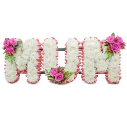 This carefully composed memorial spells MUM in three substantial white letterforms, each letter hand-filled with tightly clustered white chrysanthemums that form a seamless, pillowy surface. The chrysanthemums give a soft, subtle scent beneath the faint morning light and a tactile, linen-like texture when viewed up close. Each letter's outline is lovingly trimmed in a soft ruffled pink fabric, a tender contrast that frames the white with a blush of warmth and maternal grace. A dark green metal support is discreetly visible between the letters, the engineering behind the piece that keeps the tribute standing with quiet strength. To balance the purity of the mums, the arrangement features three small, artful clusters of fuchsia pink roses - one at the top left of the first M, one nestled in the heart of the U's curve, and one at the top right of the final M. These roses are paired with ferny green foliage and handfuls of coral hypericum berries, their lively colour and glossy texture providing focal points that draw the eye across the display. The composition reads as a peaceful, comforting homage: classic and restrained, yet intimate in its details. As a Stanmore florist familiar with services around Stanmore Broadway and Canons Park, I see this style chosen by families who want a traditional, elegant expression of love and farewell for a beloved mother.