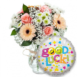 Soft morning light reveals a thoughtful Good Luck gift, its colours made vivid against a white studio background. Center-left a clear glass vase holds a lush, generously arranged bouquet: gentle light-pink roses curl inward in satin layers while sun-warm peach Gerbera daisies, dark-centred and textural, create rhythmic focal points. Dainty sprays of white baby's breath weave between stems like lace, and clusters of white-and-lime-green chrysanthemum florets add a crisp, buoyant rhythm among deep green foliage that gives the composition depth and structure. The vase's rustic twine tie suggests an artisanal touch, the sort of finishing detail a neighbourhood florist from Stanmore Common might use when packing blooms for a thoughtful walk-home gift. In the foreground, slightly to the right and overlapping the bouquet, a round metallic silver foil balloon flashes playful colour: scattered stars, hearts and confetti motifs surround bold, multi-coloured letters spelling Good Luck, with two yellow smiley faces substituting for the Os - one winking, one making a playful face - creating a warm, human moment. The metallic surface captures reflected highlights, echoing the glass vase beneath. This image smells faintly of roses and fresh greenery and reads as a confident, encouraging gesture - perfect for sending to someone facing exams, a new job, a farewell or a fresh start in life close to home in Stanmore.