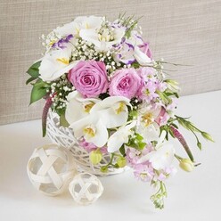 An elegant wedding-style centrepiece rests in a round white wicker basket, captured in a softly lit scene that would feel right at home on a reception table in Stanmore. The arrangement is lavish yet serene, structured around several large, fully opened pink roses, their spiral petals unfolding in gentle layers that create a romantic focal point. Interspersed among these are exquisite white Phalaenopsis orchids, their wide, porcelain-like petals spreading outwards to reveal warm yellow and soft amber throats, adding a luxurious, contemporary note. Around this central pairing, ruffled white lisianthus blooms, some kissed with a hint of lavender along their petal edges, provide delicacy and textural richness. Slender spires of pale pink stock rise and then drift outward over the rim of the basket, lending a sense of natural flow, as if they've just been gathered from a garden walk near Bentley Priory Reserve. Threaded between all the larger flowers are tiny clusters of white gypsophila, each minute bloom forming part of a misty, cloud-like layer that softens the overall silhouette. Fresh green foliage emerges here and there, giving the design structure and a lively contrast to the pastel tones. The basket, finely woven and painted white, sits on a spotless white surface, reinforcing the clean, celebratory feel. To one side rest two decorative spheres made of pale woven rattan, their open, looping bands echoing the texture of the basket and adding a contemporary, organic accent that would suit a stylish Stanmore dining room. In the background, a light beige, fabric-like surface offers gentle texture without distraction. This arrangement expresses quiet sophistication and is ideal for weddings, anniversaries, or formal dinners across Stanmore and the surrounding area.