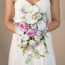 This close-up image from a Stanmore wedding tells a love story in gentle florals and pastel hues, focusing on an artful bridal bouquet that evokes the tranquillity of Springwell Avenue gardens. Pictured from the waist up, the bride holds a breathtaking cascade of blooms: the largest and most graceful are white phalaenopsis orchids, their wide petals gleaming softly and golden centers adding a sunlit touch. Nestled among them are plush, pastel-pink garden roses, their velvety petals layered with tender beauty. Subtle lavender accents bloom from clusters of stock flowers and finely ruffled lisianthus edged in violet, their spiky forms lending a hint of playful elegance. Airy sprays of baby's breath float amongst the main flowers, bringing a cloud-like softness, while just a touch of green foliage peeks through, grounding the bouquet in nature. The strapless, lace-adorned wedding dress visible below the bouquet catches the ambient light, all intricate details standing out in gentle contrast to the creamy beige backdrop-a scene reminiscent of a Stanmore morning blessed with peace and quiet joy. The bride's hands rest in a gentle curve, displaying the bouquet's flowing artistry, expertly created by a local florist attuned to Stanmore's refined tastes. Altogether, the bouquet is as much a symbol of the neighbourhood's character as it is a romantic accessory, radiating a sense of timeless love and anticipation for the celebration ahead.