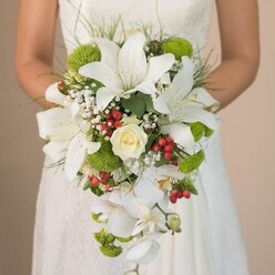 Imagine a Stanmore bride gracefully holding an opulent bouquet, the soft sunlight filtering through tall windows to illuminate each intricate bloom. At the fore, grand white lilies unfurl, their distinctive trumpet petals adorned with delicate russet speckles, invoking both purity and contemporary flair. A sumptuous cream rose anchors the centre, its coiled petals tightly nestled-a silent promise of steadfast love, resonating with Stanmore's cherished wedding customs. Trailing downward, an abundance of white Phalaenopsis orchids introduces a luxurious cascade, their gentle movement reminiscent of morning breezes along the green lanes of Stanmore. Wisps of white baby's breath scatter among the larger blooms, presenting a feathery softness akin to clouds drifting past Bentley Priory. Playful lime-green pom-pom chrysanthemums inject a cheerful, modern contrast, their hue reminiscent of the verdant local parks. Dotted throughout, clusters of glossy red Hypericum berries radiate joy and provide a striking, seasonal touch. The arrangement is expertly encircled by verdant foliage and slender grass-like greens, adding fullness and a touch of wild elegance. The bride's poised hands, holding the bouquet against the delicate lacework of her gown, bring together traditional refinement and local character. This luxurious bouquet crafted by Flower Delivery Stanmore exemplifies artistry for those seeking unforgettable wedding flowers and inspiration from the area's vibrant floral scene.