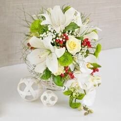 On a pristine white surface, a lavish floral display unfolds in soft whites, fresh greens, and touches of deep red, echoing the refined yet leafy character of Stanmore. The arrangement sits in a round white wicker basket, its tightly woven pattern providing a subtle rustic contrast to the polished table. From this base, several large white lilies rise confidently, their wide trumpet-shaped petals arching outward and revealing speckled interiors and golden stamens that seem to glow in the gentle indoor light. Near the crown of the bouquet, a single cream-yellow rose nestles with quiet grace, its spiralling petals velvety and full of promise, like the opening moments of a wedding reception at a Stanmore country house venue. Cascading down the right side, a stream of white phalaenopsis orchids flows in a loose teardrop shape, each orchid bloom pristine and silky, giving the entire design a bridal, almost ethereal quality. Around these focal flowers, tufts of vivid green pom-pom dianthus and an assortment of greenery, from fine, feathery foliage to broader, glossy leaves, create layers of depth and movement reminiscent of walks through Bentley Priory Nature Reserve. Tiny clusters of white gypsophila float gently between the larger blooms, while rich red berries gleam like small jewels, introducing warmth and seasonal charm. To the left of the basket, two white wicker spheres, one larger and one smaller, rest on the table, their open, interlaced design mirroring the basket and adding a carefully styled finish. The neutral beige background, with its linen-like texture, allows the crisp whites and vibrant greens to stand out. The overall mood is one of elegant celebration and serene beauty, ideal for wedding tables, anniversaries, or refined gatherings across Stanmore.