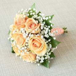 Arranged with quiet grace atop a linen-like beige backdrop, this bouquet exudes classic elegance-a favorite among Stanmore's brides and celebrants. Seven to eight full-bloomed roses, their petals tinged in nuances of peach, apricot, and pale orange, command the center with their lush, spiraling forms. The soft blush of these roses glows warmly in the kind of gentle daylight that often washes across Stanmore's charming avenues. Intertwined through the arrangement, countless white baby's breath (Gypsophila) provide ethereal contrast and a cloud-like halo, their feathery texture softening the structured round bouquet. Rich, glossy green leaves peek out from between the blooms, lending the piece both depth and a nod to Stanmore's abundant gardens. The bouquet's robust stems are carefully bound with a coral-peach satin ribbon, smooth to the touch and adorned in a spiral pattern with delicate white pearls, catching the light in a way that recalls traditional Stanmore wedding finery. The neat cut at the base of the stems highlights the freshness and purity of the arrangement. Captured from a gentle, slightly raised angle and bathed in even light, every element-from the silky petals to the luminous pearls-offers a tactile sense of anticipation and celebration. For weddings, anniversaries, or special events throughout the Stanmore area, this bouquet stands as a lasting symbol of romance, artistry, and the region's beloved floristry heritage.