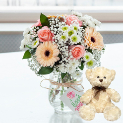 The scene shows a gentle, romantic pairing of flowers and a teddy bear, laid out as if ready to be sent for a special anniversary in Stanmore. A clear, mason jar-style glass vase holds a compact bouquet with a rounded, almost hand-tied look. Pale pink roses, with their spiralling petals, nestle beside warm peach gerbera daisies, each daisy featuring a pronounced dark brown centre that draws the eye. These standout blooms are interwoven with airy white baby's breath and small white chrysanthemums dotted with lime-green centres, creating a soft, cloud-like surround that feels both fresh and uplifting. Natural, light brown twine circles the neck of the vase and is tied with an easy bow, from which a small white gift tag hangs. On the tag, a simple pink heart design and the word Handmade in flowing green script suggest that this arrangement has been assembled with personal care, much like the bespoke bouquets crafted by Flower Delivery Stanmore for homes around Belmont Circle and Canons Park. To the right of the vase, a cream plush teddy bear sits comfortably on the same white, slightly reflective surface. Its fur appears thick and touchably soft, with dark button eyes and a small brown nose that give it a gentle, trustworthy expression. A narrow brown satin ribbon, tied neatly in a bow around its neck, adds an understated finish. The background is softly overexposed and blurred, evoking daylight streaming into a Stanmore living room overlooking local gardens or green streets. Overall, the image communicates warmth, comfort, and enduring affection, ideal for anniversaries, birthdays, or quiet thank-you moments shared across the Stanmore area.