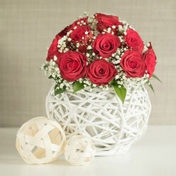 On a pristine, pale tabletop that could easily belong to a contemporary Stanmore home, a rounded bouquet of red roses becomes the undisputed focal point. The roses are a rich, velvety crimson, each bloom open and full, with petals coiling inward in neat spirals that suggest careful handling by an experienced florist. They are placed so closely together that the blossoms seem to form one continuous, luxurious surface. Threaded throughout this sea of red are airy sprays of white baby's breath, their tiny star-like flowers softening the intensity and bringing a breath of lightness to the design. Here and there, shy glimpses of green leaves offer a quiet nod to the garden, like foliage clipped from a well-kept Stanmore hedge. The bouquet rests in a distinctive white vase, bulbous and round, featuring a dramatic open-weave pattern reminiscent of twisted white branches or rattan knotted into a sculptural form. Light filters through the openings, carving gentle patterns on the table and highlighting the craftsmanship behind the piece. To the left, two decorative woven spheres in an off-white or cream tone are placed with deliberate care: the larger one nearer the front and the smaller slightly behind, their loose, ribbon-like strips forming an inviting lattice. The background is a calm, neutral wall with a soft, fabric-like texture, delicately blurred so the attention remains on the flowers and their companions. The composition speaks of romance and thoughtful detail, ideal as a wedding table centrepiece, an anniversary arrangement, or a special celebration setup for a dinner party in Stanmore with same-day delivery from a trusted local florist.