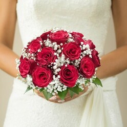 Brought to life by the expert hands of a Stanmore florist, this close-up photograph captures a bride's hands cupping a sumptuous bouquet at mid-torso level-a vision of classic wedding beauty. Dominated by deep red roses in full, velvety bloom, the design radiates both passion and refinement, their petals wrapped tightly and rich in texture. Among them, soft tufts of pure white gypsophila-baby's breath-are artfully interspersed, lending the bouquet a gentle lightness and airy contrast reminiscent of dew on Edgware's morning lawns. Sprigs of sleek green leaves peep through the blooms, a nod to the verdant spaces that grace Stanmore's landscapes. The bride's manicured fingers secure the bouquet gracefully, set against the intricate bodice of her white or ivory wedding dress, which features a delicate lace or embroidered motif extending towards a flowing sash. The softly blurred beige background directs all attention to the nuanced artistry of the bouquet and the tactile detail of bridal attire. This thoughtfully constructed image is ideal for articles on wedding trends or local Stanmore bridal inspiration, highlighting how floral craftsmanship brings romance to every celebration.