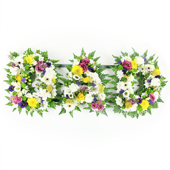 The photograph shows an elegant floral tribute that spells out "DAD" in three distinct capital letters, arranged side by side against a clean, white backdrop reminiscent of the calm interiors of Stanmore's local chapels and crematoriums. Each letter is framed by a border of deep green fern, the feathery leaves overlapping to create a soft, natural outline that sets off the flowers inside. The main body of each D and the central A is tightly packed with white chrysanthemums and large white gerbera daisies, their rounded, layered petals forming a tranquil, snowy carpet of blooms. Nestled among these whites, bright yellow gerberas and clusters of yellow spray roses offer gentle bursts of colour, their warm tones suggesting moments of joy, shared laughter, and sunlight through a window on a quiet afternoon in Stanmore. Deeper, more reflective notes come from stems of purple lisianthus and small heads of rich pink or magenta spray roses, which are placed thoughtfully so their colour feels like a soft echo of the love and respect held for a father. Here and there, petite lavender flowers and vivid green button chrysanthemums break up the surface with subtle detail, bringing texture and liveliness without disturbing the arrangement's calm dignity. The three floral letters are anchored along the bottom by slender dark green bars that keep them in perfect alignment while remaining understated. Crafted with the care of an experienced local florist, this tribute would sit beautifully at the front of a funeral service, along a hearse, or by a graveside around Stanmore, offering a tender, visually powerful farewell to a cherished dad.