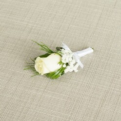 A refined wedding boutonniere is displayed on a diagonal across a pale beige, linen-like fabric, the subtle weave of the material creating a gentle backdrop that feels very much in keeping with the elegant, residential character of Stanmore. At the centre of the composition rests a single ivory-white rosebud, its petals soft and velvety, cupped close yet beginning to unfurl as if just entering full bloom. The rose is encircled by airy sprigs of white baby's breath, each tiny flower forming part of a mist-like collar around the central bloom, adding lightness and texture without overwhelming the simplicity of the design. Slender, glossy deep green leaves and a few feathery green fronds extend from behind the rose, providing contrast and structure, echoing the greenery found in local gardens around Stanmore Hill and the quiet streets off The Broadway. The stems of the rose and foliage are carefully bound together in a smooth white satin ribbon, wrapped with even precision that hints at an experienced florist's steady hands. Just below the flower head, the ribbon is fashioned into a dainty bow, its loops neat and understated, ready to sit flush against a lapel on a tailored suit or tuxedo. Gentle, diffused lighting glides over the petals and leaves, picking up subtle shadows within the rose and the fine silhouettes of the gypsophila stems. The overall mood is calm, romantic, and timeless, ideal for a groom or groomsman preparing for a wedding at a local Stanmore venue, and the image reflects the type of classic boutonniere chosen by couples who appreciate simple beauty, tradition, and careful attention to detail in their floral styling.