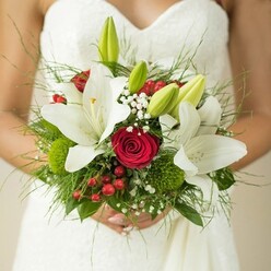 Artful and evocative, this Stanmore bridal bouquet speaks as much of skilled craftsmanship as it does of emotion. Pristine white lilies, their broad petals fully open, set a tone of innocence and clarity that mirrors the tranquil ambiance of Stanmore's suburban gardens. Rich crimson roses coil quietly among the lilies, their vibrant hue serving as the foundation for the bouquet's dynamic energy and heartfelt romance. Cheerful red berries and playful, mossy button-like greens add elements of intrigue and tactile richness, while sprigs of delicate gypsophila float throughout, reminiscent of light Stanmore breezes brushing through Bentley Priory's meadows. The varied foliage-feathery and robust-adds both depth and an organic flow, echoing the contours of parkland paths. The bride's gently clasped hands, soft with anticipation, cradle the composition as if holding hope itself. Every stem has been selected with attention to texture, colour, and longevity, making this bouquet more than a simple accessory-it serves as a cherished keepsake, a fragrant symbol of commitment and joy inspired by Stanmore's own charm.