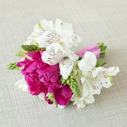 A small yet luxurious floral piece is laid out horizontally on a light, sand-coloured textile that resembles finely woven linen, its soft texture providing a subtle contrast to the crisp forms of the flowers. In the centre rests a stunning fuchsia peony, fully open and plush, with layer upon layer of ruffled petals that fold into one another like silk. This vivid focal bloom is surrounded by white alstroemeria, their slender petals slightly curved back, each one etched with fine maroon lines and speckles that add a sense of delicacy and craftsmanship. Threaded between the white and fuchsia tones are slender stems of fresh green buds and spiky clusters, lending a clean, linear texture that makes the composition feel modern while still romantic. The stems sit tightly bound with a bright pink satin ribbon, wrapped with precision and finished without visible pins or tape, giving the arrangement a polished, boutique feel that you might expect from an experienced florist along Church Road in Stanmore. The lighting is soft and even, highlighting the gentle sheen on the ribbon, the velvety surface of the peony petals, and the faint shadows that fall across the neutral background. This compact design feels like a cherished detail from a wedding or prom-a boutonniere or mini posy that could be worn at a ceremony in a Stanmore church or carried for photos in a nearby park, its colours symbolising joy, affection, and the sweetness of early summer. The overall impression is of thoughtful, effortless beauty, captured in a small handful of fresh blooms.