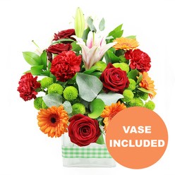Captured in bright, even light against a white background, this arrangement presents a joyful mix of colours and textures in a low rectangular glass vase encircled with a soft green and white gingham ribbon, a detail that brings to mind the gentle, suburban charm of Stanmore's tree-lined lanes. The bouquet feels plentiful and uplifting, its flowers gathered closely together in a painterly composition. Deep red roses, with densely layered, velvety petals, offer a classic romantic note, while nearby red and red-orange carnations bring softness and volume with their delicately frilled edges. Interspersed throughout, bold orange gerbera daisies with dark central disks stand like small, sunlit faces, their clean, symmetrical petals radiating vibrancy reminiscent of a sunny afternoon in Stanmore Country Park. Counterbalancing these warm tones, clusters of lime-green button chrysanthemums appear as small, round cushions of colour, adding a crisp, modern twist and a lively sense of rhythm to the design. Rising from within the bouquet, white Oriental lilies add height and elegance; several blooms are open wide, revealing subtle pink stripes and golden stamens, while tall buds wait to open over the coming days, extending the life and changing character of the display once delivered. Around the blooms, a generous collar of glossy dark green foliage and fine, silver-green eucalyptus leaves gives structure, depth, and a natural, garden-inspired feel, as if the flowers were freshly cut from a well-tended Stanmore garden. Through the clear glass, the submerged stems create a clean, linear counterpoint to the softness above. In the bottom right corner of the image, a bright orange circular label reads VASE INCLUDED in bold white capitals, indicating that this vibrant bouquet is supplied complete and ready to place straight onto a table or reception desk.