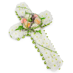 This image shows an elegant funeral flower cross arranged with the calm, measured artistry you might expect from a long-established Stanmore florist preparing for a church or chapel service. The cross form is completely covered in pristine white chrysanthemums, or similarly small white blooms, which have been placed very close together so that almost no stem or greenery shows through. The result is a soft, pillow-like field of petals that communicates purity, innocence, and serene farewell. Encircling the entire shape is a narrow border of vivid green foliage, providing a crisp frame and lending the cross a clearly defined outline, much like the manicured edges of lawns around Stanmore Country Park. Where the horizontal and vertical arms meet, a raised central spray of larger blooms creates a focal point. Several full-headed roses in shades of apricot-peach, delicate blush pink, and creamy ivory rise above the white bed, their spiralled petals gently opening as though in a quiet gesture of love. These roses are nestled among lacy fern fronds, fine arcs of bright green grass that loop and sweep, and tiny touches of purple filler flowers which bring subtle colour and depth without overwhelming the overall white theme. The lighting is even and bright, allowing the textures and layered details of each bloom to be seen with clarity. This traditional Christian cross arrangement is most often chosen for funerals and memorials in and around Stanmore, perhaps gracing the front of a church near Stanmore station or resting by the chapel at a local cemetery. It speaks softly of faith, remembrance, and the hope of eternal peace, offering a graceful floral expression of sympathy to those who are mourning.