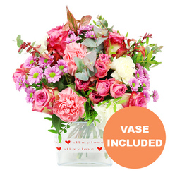 A bright, romantic bouquet fills the centre of the frame, overflowing from a clear, square glass vase that stands on a pure white surface against a matching white background. The light in the photograph is soft and even, allowing each flower head to show its true colour, much like a carefully composed display in a Stanmore florist's window. Large pink roses are the stars, some in deep fuchsia with slightly darker outer petals, others in gentle, even blush tones, all fully open so their layered centres are visible. These are nestled closely with generous carnations in coral, peachy pink, and creamy white, the ruffled edges giving the arrangement its sumptuous volume and a tactile richness you can almost feel. Threaded throughout are smaller daisy-like chrysanthemums in shades of lavender and mid-purple, their round, lime-green centres creating a crisp contrast that enlivens the whole design. The bouquet is cocooned in foliage: rounded eucalyptus leaves with a cool, silvery-green tone, alongside deeper green sprigs and hints of reddish-brown leaves that add depth and a subtle nod to autumn. The stems are clearly visible through the glass, fanning out in clean, green lines. Wrapped around the lower part of the vase is a white decorative band printed with tiny red hearts and the affectionate script all my love, reinforcing the sense that this is a heartfelt, personal gift. In the bottom right corner, a bold orange circular overlay with clear white capital letters reads VASE INCLUDED, indicating that the arrangement arrives exactly as shown, ready to be placed on a dining table in a Stanmore home or gifted for a special occasion. The overall feel is of a joyful, loving bouquet, ideal for anniversaries, Mother's Day, or simply brightening someone's week.