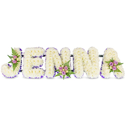 There is an understated artistry in this name tribute: the five letters composing JENNA emerge as soft, sculpted forms crafted from a mosaic of small, snowy-white chrysanthemums or like blooms. The dense flowering gives each letter a pillowy depth, a tactile softness that contrasts gently with the clean white background. A delicate stroke of purple - either a thin ribbon or tiny lavender florets - outlines the inner edges and base of each block letter, providing a refined border that reads like a quiet frame. Intermittent clusters of spray roses in blush-pink and muted lavender, finished with a few cream or pale yellow buds, are carefully nested into three letters to draw the eye and introduce intimate points of colour. Dark green fern fronds fan around these clusters, offering slender, feathery movement against the compact floral mass and suggesting a woodland freshness. The composition rests on slim, dark connecting bars, implying secure construction for display in church aisles or at a funeral reception table. Light is soft and even, picking out subtle shadows and the gentle sheen of petals, evoking memory and the hush of reflection. This floral lettering reads as a handcrafted salute - a personal, name-led composition often chosen by families across Stanmore and nearby Harrow when they want a dignified, bespoke tribute. The overall effect is one of quiet elegance: grief held with care, beauty chosen as a language of remembrance.