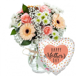This photograph portrays a thoughtful Mother's Day gift set, styled as though ready to be delivered to a quiet home in Stanmore, perhaps just off Uxbridge Road or near the green spaces of Canons Park. The composition is clean and bright, with every element standing clearly against a white background. On the left rests a clear, straight-sided glass vase, its transparency revealing the gathered stems within. Around the neck, a length of light brown twine is tied in a simple bow, adding a touch of rustic personality that softens the formality of the glass. Rising from the vase is a lush, rounded bouquet of pastel flowers. Several peach-pink roses open in generous spirals, their petals layered like soft fabric. Large peach gerbera daisies punctuate the bouquet, their daisy-like petals framing bold, dark brown centres that provide a focal point. Intermingled among them are small white chrysanthemums with pale green cores and airy clusters of white baby's breath, which appear almost like fine, floating bubbles of light. Fresh green foliage threads through the blooms, framing the colours and suggesting the crisp scent of newly cut stems. Behind the bouquet, almost hidden, is a small white greeting card with hints of green and pink handwriting, bringing a personal whisper of "Happy Mother's Day" from child to mother. To the right of the vase a heart-shaped foil balloon stands proudly forward. Its glossy white surface is dotted with sparkling gold and silver spots, and in the middle a soft peach heart holds the words "HAPPY Mother's DAY" written in bold black script, the word "Mother's" drawn in graceful, calligraphic strokes. The overall impression is of joy, tenderness and gratitude, ideal for same-day Mother's Day flower delivery across Stanmore and neighbouring Harrow Weald.