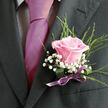 The image presents a detailed close-up of a gentleman's formal attire, captured from the shoulders to mid-chest, as if he were about to step into a wedding ceremony or black-tie celebration in Stanmore. The suit jacket is a rich charcoal grey, tailored cleanly so the lapel lines are sharp yet understated, the fabric absorbing the light in a way that makes the colours of the flowers and tie stand out. A pale dress shirt with a small check or gingham pattern lies beneath, its neat collar framing a smooth, vibrant magenta tie that falls in a confident line down the centre of the image. The tie's satiny surface reflects a gentle sheen, hinting at late-afternoon light before an event. Pinned securely to the left lapel is a single, exquisite light pink rose, its petals unfurling in soft layers from a deeper, rosy core to a delicately blushed edge. This rose sits at the heart of the boutonniere, surrounded by tiny white gypsophila blossoms, their lacy clusters giving a clouded, romantic softness to the arrangement. Fine strands of green foliage flick upwards and outwards, adding movement, as though they were freshly cut from a garden near Canons Park. At the base, a broader green leaf or band holds the stems, neatly finished with a slim, glossy purple ribbon tied into a modest bow. The colours of rose, ribbon and tie harmonise quietly, conveying affection and intention without overpowering the simplicity of the suit. The whole scene suggests a poised, hopeful moment just before vows are exchanged or a milestone celebration begins in Stanmore's cosy, familiar streets.