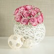 A softly lit indoor scene shows a rounded bouquet of blush pink roses and airy white baby's breath resting in a spherical woven white vase, styled as if for a calm afternoon in a Stanmore living room. Around fifteen to twenty roses are clustered closely together, each bloom opening generously, with petals that begin as a delicate, almost porcelain pale pink at the centre and deepen to a richer, near-fuchsia tone at the edges. The roses are laced throughout with fine sprays of Gypsophila, their tiny white blossoms and slender green stems creating an almost cloud-like halo that lightens the arrangement and adds a sense of romance and gentleness. The flowers sit neatly within a round, basket-style container crafted from white, interlaced fibres, reminiscent of painted wicker or rattan. Its open criss-cross design allows slivers of light to pass through, casting subtle shadows on the smooth white surface below. In front and to the side of the vase rest two decorative spheres made from pale, natural wood strips, woven into open geometric balls of different sizes, echoing the organic textures of the vase. Behind, a softly textured wall in warm beige or cream offers a quiet, neutral backdrop, allowing the pinks and whites to glow without distraction. The overall composition feels serene and contemporary, with a hint of handmade charm often seen in carefully styled homes around Stanmore, from tidy terraces off Church Road to bright flats near Stanmore station. It is the kind of delicate floral display you might choose for a bedside table, a thoughtful anniversary gift, or a gentle welcome arrangement for guests arriving on a Sunday afternoon.