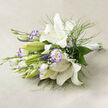 On a soft, neutral linen surface, arranged horizontally and photographed in gentle daylight, this bouquet presents a fresh, early-summer temperament. The scene is led by pristine white lilies, their trumpet-shaped faces opening boldly to reveal luminous, creamy throats, warm golden stamens and the tiny dark flecks that give each blossom character. Alongside them, several green buds sit reserved and expectant, promising stages of bloom to come. White lisianthus weave through the lilies; their ruffled petals bear a subtle lavender-purple wash or defined edge that creates a delicate, almost watercolor contrast against the dominant whites. Fine, feathery sprigs of foliage-soft as breath-move through the arrangement, while larger, deeper green leaves anchor the mass and add a reassuring weight.

Tiny clusters of baby's breath are woven through the bouquet like threads of mist, softening transitions and enhancing the airy composition. The stems are gathered to the right and secured with a crisp white satin ribbon, the tidy hand-tied finish lending the piece a modern yet timeless silhouette. Natural lighting sculpts soft shadows across the linen, allowing textures to read clearly and highlighting the florist's intent to balance fullness with lightness.

This bouquet carries a calming, restorative energy: suited to a serene gathering, a thoughtful gift, or as an elegant wedding choice. It communicates refinement without fuss, the work of someone attentive to proportion, scent and the tactile joy of fresh blooms.
