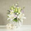 A calm, sophisticated floral arrangement featuring white lilies and lisianthus is displayed in front of a soft beige backdrop, capturing the understated elegance often favoured for weddings and sympathy flowers in Stanmore. The bouquet is composed on a spotless white surface, illuminated by clear studio light that sharpens every edge yet keeps the mood gentle. At its heart, large white lilies are fully open, their petals arching outward in smooth, slightly curled layers that expose finely speckled centres and prominent yellow-orange stamens dusted with pollen. Encircling these focal blooms, lisianthus flowers in shades of white and pale lavender add softness; some petals are tightly cupped, others forming delicate ruffles with faint violet outlines, like watercolour strokes. Slender stems of dark green foliage and airy clusters of tiny white filler flowers, similar to baby's breath, weave between the lilies and lisianthus, creating depth and a sense of light drifting through the design. A few unopened lily buds in pale green emerge from the top, suggesting the piece will continue to evolve, just as celebrations unfold in homes near Stanmore Country Park or along The Broadway. The bouquet is held in a round white vase made of finely interlaced strands, producing a basket-like lattice that feels modern and sculptural. Beside the vase, two rattan-like spheres in a natural beige tone echo its texture, grounding the cool whites with an earthy warmth. Behind, a subtly textured beige background with fine vertical lines adds a refined, woven effect without distracting from the flowers. The image conveys quiet grace and emotional reassurance, well-suited for wedding collections, condolence arrangements, or minimalist interior styling across Stanmore.