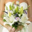 Like a breeze over Stanmore's meadows, this wedding bouquet whispers of hopeful beginnings and refined beauty. In the bride's softly polished hands nestle velvety white Oriental lilies, their petals opening to reveal jeweled yellow stamens, each flower brimming with pure, serene fragrance. Among them, lisianthus provide a gentle contrast-ruffled white blooms edged in delicate lavender, intermingling with unopened buds that suggest stories yet to unfold. Pops of striking purple, likely Veronica, lend height and vibrancy, while feathery baby's breath floats between the blooms, casting a cloud-like magic over the entire bouquet. Surrounding it all, a tapestry of lush green leaves and wispy asparagus ferns imparts a feeling of wild grace, recalling walks through Stanmore's leafy parks and gardens. Hints of the bride's strapless gown blur softly in the background, making the florals the star of the scene. Careful, loving craftsmanship is evident in every detail-a bouquet that is not only beautiful to behold, but carries the soul of a Stanmore wedding day within its blooms.