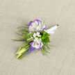 A compact study in spring restraint, this boutonniere pairs two cool, ruffled lisianthus blooms-pure white with a whisper of violet along their edges-with clouds of tiny white baby's breath and pale, slender buds that suggest gentle movement. Below them, short clusters of grape hyacinth give a concentrated flash of blue-violet, their upright spikes introducing texture and visual punctuation, while filigree ferns fan around the composition to add an airy, woodland silhouette. The stems are neatly wrapped in white satin ribbon, the knot pressed flat on neutral linen that frames the piece and highlights the interplay of soft fibres and velvety petals. In low, diffuse daylight the colours remain demure yet distinct: the violet edging reads almost iridescent against the clear white, and the hyacinth's deeper blues anchor the palette. There is a faint, pleasing scent when the boutonniere is held close, a whisper of floral sweetness that suits intimate occasions: weddings on Stanmore High Street, anniversary portraits near Bentley Priory, or a formal family photograph on a crisp spring afternoon. Prepared with care in our local workshop, Flower Delivery Stanmore arranges each stem to sit comfortably on a lapel, mindful of balance, scale, and the tactile pleasure of silk-soft petals against wool or linen. The piece feels both contemporary and quietly traditional-a small, intentional keepsake that reads as elegance in miniature and carries the memory of the day in its temperate hues and thoughtful construction.