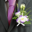 This boutonnière is a study in restrained elegance, crafted to sit just above the heart and to be noticed without shouting. The centrepiece is a white lisianthus, its silky petals rimmed with a bold purple edge that defines each curl and adds instant drama. A compact white dianthus with lively purple ruffles nestles close, its serrated texture providing a lively contrast to the lisianthus' smoothness. Behind them three pale-green lisianthus buds point upward, a small promise of blooms to come, while a slim stem of deep purple veronica extends like a composer's vertical accent. Lush, dark-green foliage and looping ornamental grass weave through the arrangement, introducing soft movement and a natural, garden-fresh feel; tiny sprays of gypsophila scatter like breathy clouds, lending lightness and a whisper of innocence. All elements are tied with a crisp white ribbon, finished to lie flat and secure against the lapel of a charcoal suit and to complement a lustrous magenta tie. Morning light picks out the purple rim and the velvety petal surfaces, and the scent is mild-green, slightly sweet, reassuring across long hours of celebration. Prepared in our Stanmore studio and chosen often by couples planning ceremonies near Bentley Priory or deliveries to houses around Stanmore Underground, this boutonnière balances tradition with personality. Flower Delivery Stanmore packages it to travel safely and arrive fresh, a thoughtful accent for grooms, best men, or any guest whose detail matters on a meaningful day.