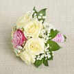 A dainty hand-tied bouquet is shown laid diagonally on a softly textured, light beige fabric, whose fine, natural-looking weave evokes the feel of linen napkins on a carefully dressed Stanmore dinner table. The arrangement centres on five creamy white roses, each bloom full and rounded, with silky petals that curl outward in gentle, layered waves, suggesting freshness and an understated elegance. Nestled close to the front-left is a single, more vivid rose in bright fuchsia pink, its rich colour bringing a playful spark and focal point to the otherwise gentle palette. Between and around the roses, clouds of delicate white baby's breath weave in and out, their tiny blossoms forming a light, lacy veil that softens the transitions between ivory and pink. Deep green, pointed foliage fans out around the flowers, offering a grounding contrast and a touch of the garden, as if freshly gathered from a well-kept Stanmore back garden near Stanmore Hill. The stems are gathered tightly and wrapped with a lustrous, bright pink satin ribbon, creating a smooth, tailored handle that looks comfortable to hold for long celebrations. Over this ribbon, fine strands of small, iridescent pearls are aligned in parallel horizontal rows, adding a sense of ceremony and careful craftsmanship. The soft, even lighting in the image highlights the natural curves of each petal and leaf without harsh shadows, preserving a calm, romantic atmosphere. This petite arrangement feels ideal as a corsage, boutonnière, or an intimate centerpiece for weddings, proms, or engagement dinners in Stanmore, embodying a subtle blend of tradition, joy, and polished style.
