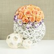 On a smooth white tabletop that reflects the faintest outline of its shapes, a refined floral arrangement radiates gentle warmth, as if awaiting guests at a Stanmore wedding breakfast or family celebration. The centerpiece rises in a generous dome of roses in peach, apricot, and soft salmon tones, each bloom fully open and nestled tightly against its neighbours. The layered petals are velvety and full, creating a rounded, cushion-like profile with subtle tonal variations that move from pale outer petals to richer, glowing centres. Around the base of this rose sphere, a graceful fringe of white lisianthus encircles the arrangement. Each lisianthus bloom is edged with a bold band of violet and purple, adding a striking yet harmonious contrast that echoes the kind of careful colour pairing a local florist at Flower Delivery Stanmore might create. Between these blooms, tiny sprays of white baby's breath weave in and out, their starry flowers adding a weightless, cloud-like softness. The flowers are arranged in a distinctive, bulbous white container woven from slender wicker or rattan strips. This open lattice structure forms a broadly spherical shape, allowing the neutral background to show through and creating an interplay of light and shadow that feels modern but still rooted in natural craft. Resting on the tabletop in front of the arrangement are two decorative spheres, one larger and one smaller, crafted from pale, natural wood-coloured strips twisted into airy orbs. Their texture mirrors the vase and lends a calm, sculptural balance to the scene. The warm, subtly textured beige backdrop frames everything with understated elegance, making this arrangement ideal for wedding décor inspiration, sophisticated table styling, or serene home interiors across Stanmore.