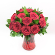 A studio-style image of a classic romantic bouquet shows a generous cluster of deep red roses arranged in a rounded, almost perfectly symmetrical dome, set sharply against a pure white background. Each rose is a rich crimson, with velvety petals that curl inward toward tightly furled centres, giving the blooms a sense of poised anticipation, as if they are just about to open further. Around and between the blossoms, glossy dark green leaves, likely Ruscus, peek through in carefully spaced intervals, softening the lines of the bouquet and creating a vivid contrast that makes the red appear even more intense. The foliage gathers at the base, framing the flowers and lending the arrangement a sense of natural fullness and depth. All of this is gathered into a translucent ruby-red glass vase with a subtle swirling or ribbed texture that catches the light in delicate highlights and shadows. Through the coloured glass, the rose stems are visible as vertical, slightly crossing lines, adding organic detail beneath the blooms. The vase has a gently flared top and base, giving it a stable, elegant silhouette that supports the drama of the roses above. Bright, even lighting highlights every curve of petal and sheen of leaf, making the arrangement look freshly made, as if it has just left the workbench of an expert florist in Stanmore. The overall mood is one of passionate romance and confident affection, perfect for a Valentine's Day surprise, a milestone anniversary, or an unforgettable declaration of love delivered to a home near Stanmore Hill or along The Broadway.