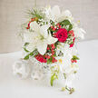 In this refined wedding arrangement, creamy white lilies and classic red roses are gathered into a softly rounded bouquet that looks entirely at home in a Stanmore reception venue or a sunlit dining room near Canons Park. The lilies sit proudly at the heart of the display, their broad ivory petals opening like calm, star-shaped fans, each bloom edged with a faint greenish tint that highlights their freshness. Between them, lush red roses nestle close, their petals layered into deep spirals of colour that read as both passionate and reassuring, like a long, steady love. Wisps of gypsophila thread their way through the larger flowers, forming delicate clouds of tiny white blossoms that soften the strong contrast of red and cream. Scattered clusters of hypericum berries add pops of glossy, coral-red brightness, reminiscent of small jewels or berries glimpsed on a walk around Stanmore Country Park. Gentle greenery and fine stems help guide the eye in an easy circle, giving the bouquet a natural, garden-inspired rhythm rather than a strict, formal structure. The flowers rest inside a white woven container whose simple, basket-like texture gives a subtle rustic note while keeping the overall look polished and elegant. Imagined in the soft light of a wedding breakfast, the bouquet seems to breathe with life: the cool silkiness of lily petals, the velvet touch of rose heads, and the airy whisper of baby's breath working together to evoke tenderness, joy, and a sense of occasion. It is a design that balances tradition with contemporary ease, perfectly suited to celebrate heartfelt milestones across Stanmore and the surrounding neighbourhoods.