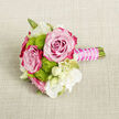 A delicately crafted mini-bouquet lies on a softly textured, light beige fabric, the neutral background allowing each flower to stand out with clarity and grace. At its heart, two lush roses unfurl in full bloom, their petals graduating from tender pastel pink in the centres to a more vivid fuchsia at the tips, giving the impression of hand-painted colour gradients often seen in carefully cultivated garden roses around Stanmore's leafy streets. Between these statement blooms, bright lime-green button chrysanthemums cluster in compact domes, each tiny floret adding depth and a playful, tactile quality, like cushions of fresh spring grass. Encircling this core are small white flowers and creamy buds, close in form to lisianthus or freesia, some fully open with narrow petals and soft yellow-green eyes, others still gently closed, promising to unfold. Fine green stems and slender foliage lend subtle structure, without overwhelming the petite scale. The stems are neatly gathered and encased in a vivid fuchsia satin ribbon, smooth and slightly glossy, echoing the roses' deeper tones. Winding elegantly around this handle is a single strand of pearl-like beads, dainty and lustrous, adding a bridal touch that suggests weddings at venues near Bentley Priory Museum or intimate celebrations in Stanmore homes. The overall composition feels fresh, romantic, and carefully considered, perfectly suited as a groom's or usher's boutonnière, a corsage, or a small bridesmaid posy, capturing the scent and feeling of a calm spring day, where subtle rose fragrance mingles with crisp greenery and quiet anticipation.