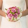 In Stanmore's gentle afternoon glow, a bride's bouquet becomes a symbol of romance and locality-an intricate composition held delicately against the refined background of an off-white lace gown. The eye is instantly drawn to the lush, round roses at the core of the bouquet, each unfurling in pastel lavender and dusty mauve, their edges kissed by bold strokes of fuchsia. These roses nestle alongside plump lime-green chrysanthemums that sparkle with freshness and texture, mirroring the vibrancy of Stanmore's community gardens in peak bloom. The bouquet's circumference is woven with clusters of petite, immaculate white blooms, most likely elegant spray roses or sweet-smelling freesia, their subtle scent echoing the fragrance of a garden after summer rain. Throughout, tiny green shoots and leaves lend fullness and introduce natural whimsy. The bride's soft pink nails, featuring a single, sparkling detail, clasp the bouquet with grace-capturing a candid moment of anticipation unique to Stanmore weddings. The scene is softly illuminated, accentuating the layers of lace, multifaceted blooms, and the tender artistry behind each floral choice. Created by experienced Stanmore florists, every element in this bouquet tells a story of love, renewal, and the rich traditions cherished in this inviting North London locale.