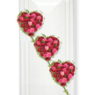 A quietly powerful image shows three heart-shaped floral tributes arranged in a gentle diagonal, each one crafted entirely from red and pink roses. The hearts are full and rounded, made by packing the blooms so closely together that they form a seamless field of petals, from deep crimson to vivid fuchsia and soft blush. The darker roses add a sense of depth and solemn love, while the pink shades bring warmth, compassion, and a feeling of tender remembrance. Among the flowers, subtle differences in bloom stages-from tight buds to wide, open blossoms-give the design texture and a sense of life unfolding. Each heart is edged with a graceful frame of fresh ivy, the fine stems curving along the lines of the hearts and linked to one another in a slender chain of glossy green leaves. This delicate greenery connects the three shapes visually and symbolically, suggesting the continuity of love across time and between generations. The arrangement appears against a pristine white background, similar to a smooth wall in a Stanmore funeral chapel or a calm room near Stanmore station, where there is nothing to distract from the colours and shapes of the flowers. Soft, even lighting highlights the velvety nature of the petals and the gentle shine of the leaves, casting a faint shadow that hints at the depth of the piece. This tribute feels both romantic and respectful, ideal for a final farewell that honours a loved one with beauty, sincerity, and quiet grace.