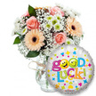 A cheerful Good Luck gift set captured against a clean white backdrop: a generous posy of soft pastel blooms sits slightly left in a clear glass vase, its neck finished with a rustic twine bow that hints at hand-tied craftsmanship. The arrangement reads gentle and optimistic - powder pink roses with tightly furled petals nestle among warm peachy-orange Gerbera daisies whose dark brown centres give them a vintage, tactile warmth. Fine sprays of delicate white baby's breath (Gypsophila) drift like lace through the composition, while small white and lime-green chrysanthemum florets add bright pops of contrast against deep glossy foliage. The flowers carry a subtle layered scent, the rose's soft perfume balanced by the fresh, clean note of the chrysanthemums and the faint, leafy earthiness of the greenery. To the right and overlapping the lower portion of the vase is a round silver foil balloon that gleams with metallic sheen; its surface is patterned with playful hearts, stars and tiny multicoloured confetti shapes, and the large, bubbly Good Luck text is painted in lively rainbow tones. The first word is punctuated by two sunlit yellow smiley faces - one winking, one sticking its tongue out - lending a familiar, encouraging cheer. At the balloon's base a small brand logo is visible, grounding the scene in real-world detail. This composition feels like a friendly send-off from a local Stanmore table - the kind of uplifting arrangement a neighbour might leave at Stanmore Broadway to share encouragement for exams, interviews, or a new beginning.