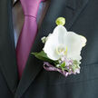 In this image, an elegant wedding boutonniere rests just above the pocket of a dark, finely cut suit, evoking the polished calm of a Stanmore ceremony. The focal point is a single white orchid, luminous and serene, its petals unfolding in smooth, rounded layers. Each petal has the softness of fine china, with a faint ivory wash along the edges that keeps the white from feeling stark. At the centre, a gently shaded throat adds an intriguing depth. Around the orchid, slim, cool-toned green leaves curve in a protective frame, while slender stems of lilac blooms and tiny white filler flowers thread through, adding lightness and movement. These lilac details suggest a soft, floral scent-subtle, not overpowering-like the air drifting through gardens off Marsh Lane on a spring morning. The stems of the boutonniere are neatly wrapped in a narrow violet ribbon, its satiny texture catching small glints of light, and the shade harmonising perfectly with the lilac tie and crisp shirt just visible beneath the lapel. The rich charcoal cloth of the suit creates a strong contrast, so the orchid seems almost to hover above the pocket, glowing slightly under the gentle lighting. The design feels carefully considered and unmistakably romantic, the sort of finishing touch chosen by couples celebrating at venues around Stanmore Hill or Bentley Priory Museum. Every element-flower, foliage, and fabric-works together to express quiet confidence, love, and the expectant happiness that lingers in the air just before the vows are spoken.