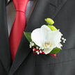 This close-up image reveals a carefully arranged boutonnière pinned to a black suit jacket, set against the polished richness of a red tie, evoking the atmosphere of a wedding day in Stanmore. The focal bloom is a white orchid, its petals broad, velvety, and gently curved, arranged with an almost sculptural symmetry. At the heart of the orchid, a subtle hint of yellow glows softly, giving the flower depth and an inviting warmth. Just above and behind it, lime-green button chrysanthemums form compact, spherical blooms, their fine, densely layered petals adding a lively, tactile texture, like tiny bursts of fresh greenery. Threaded through the design are airy sprigs of baby's breath, their minuscule white blossoms scattered like stars, lending a light, misty quality that softens the stronger shapes. Beneath the flowers, glossy deep-green foliage fans outward, framing the composition and echoing the vitality of Stanmore's leafy residential streets and parks. The stems of the boutonnière are neatly bound with a slender strip of dark red ribbon, echoing the tone of the tie beneath and introducing a subtle note of passion and romance. The interplay of colours-white, green, and crimson-creates a striking contrast against the dark suit, yet the overall mood remains graceful and composed. One can almost picture the wearer standing outside a Stanmore registry office or local venue, the fresh flowers catching the natural daylight during photographs. This boutonnière feels intentionally personal, a small but significant touch chosen to reflect taste, commitment, and attention to detail, designed to stay fresh and secure from the walk down the aisle to the final farewell at the end of a joyful celebration.