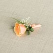 A softly lit close-up shows a meticulously crafted peach rose boutonniere resting horizontally on a pale beige linen-style fabric, the fine weave of the material forming a calm, neutral backdrop. At the heart of the arrangement sits a single rose in partial bloom, its petals spiralling outward from a warm, apricot-peach centre to creamy, almost vanilla-toned edges, giving the impression of a flower just at its peak of freshness. Nestled around the upper edge of the rose is a delicate halo of white baby's breath, each tiny blossom like a fleck of soft foam, adding lightness and a gentle, romantic texture. Slender, grass-like fronds of vivid green foliage and a couple of slightly broader, deeper green leaves emerge from behind the rose, subtly curled to cradle the bloom and give the boutonniere a natural, sculpted shape. The visible stems are neatly bound in a matte peach ribbon that closely matches the rose, wrapped with careful tension and finished in a small, tidy bow near the base, suggesting a florist's practiced hand. Soft, even lighting catches the satin sheen of the ribbon, the velvety surface of the petals, and the crisp edges of the linen, creating a serene, almost timeless mood. The overall composition suggests a refined wedding or prom accessory, perhaps waiting to be pinned to a lapel before a ceremony at a church or venue in Stanmore, close to leafy parks and quiet residential streets, where understated floral details like this help tell the story of a tender, memorable day.