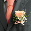 Pinned with careful precision to the left lapel of a deep slate suit jacket, a single peach-pink rose becomes the intimate focal point of an otherwise structured outfit. Each petal is shown with soft, layered edges that are just starting to unfurl, and the blossom sits in a pool of gentle daylight that reveals subtle veining and a polished, velvety surface. Surrounding the rose, delicate sprays of white baby's breath (gypsophila) float like tiny stars, while fine green tendrils weave a lacy framework that softens the jacket's tailored silhouette. The boutonnière is finished with a slender pale-pink satin ribbon tied in a small, tidy bow, its sheen reflecting the warm tone of the matching peach tie worn over a light-coloured shirt patterned with a faint dark grid. The contrast between the soft, fragrant bloom and the crisp, pronounced lines of the charcoal fabric creates a feeling both romantic and refined-perfect for a wedding, anniversary, or formal celebration. As a florist who knows Stanmore and its corners from Stanmore Broadway to Stanmore Park Road, I imagine preparing this piece in the calm before a ceremony, perhaps collected by a groom heading to the local church or to a reception near Stanmore Common. Flower Delivery Stanmore often crafts boutonnieres with this same attention to balance and scale, choosing blooms and foliage that will sit comfortably against tailored menswear while offering a quiet scent of garden-summer and a visible note of care.
