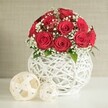 A romantic floral display is arranged on a smooth, pale surface, reminiscent of a carefully laid wedding table in a Stanmore home overlooking leafy streets near Stanmore Country Park. At the centre stands a striking bouquet of deep crimson roses, gathered into a rounded dome that overflows with velvety petals. Each rose appears freshly opened, the inner spirals still tight while the outer petals curl gently away, creating lush, layered textures. Threaded among the roses are fine clusters of white baby's breath, their tiny blossoms forming a delicate mist around the richer tones of red, bringing lightness and a sense of air between the blooms. Subtle touches of green foliage flicker at the base, adding a natural accent without distracting from the main composition. The bouquet is housed in an elegant round vase, pure white and sculptural, with an open-weave structure that looks as though slender white branches have been intertwined into a hollow sphere. This airy design allows glimpses through to the background and lets soft light pour through, casting gentle shadows and highlighting the sculptural nature of the container. To the left of the vase, two decorative spheres made from flat, cream-coloured strips are casually placed, one larger orb closer to the viewer and a smaller one tucked behind. Their open lattice design echoes the texture of the vase and introduces a relaxed, contemporary feel. Behind it all, a subtly textured, beige-toned wall provides a calm, neutral backdrop that keeps the focus firmly on the interplay of red, white, and green. The overall mood is one of refined romance, perfect for an intimate dinner, anniversary toast, or wedding celebration in Stanmore.
