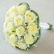 There is a serene, almost early-morning quality to this bridal bouquet: a clustered dome of around fifteen to twenty creamy ivory roses, their petals spiraled toward perfect centers, creating a plush, generous silhouette that reads as both classic and intimate. Interspersed throughout are fine sprays of Gypsophila-baby's breath-that bring a feathery, cloud-like lightness and a delicate white contrast to the fuller rose heads. Woven through the edges, flexible strands of bright green grass (bear grass or lily grass) arch in slender loops, giving the composition a modern sculptural edge and framing the blooms with natural movement. At the base, the green stems are visible and carefully gathered beneath a smooth white satin ribbon tied with a tidy knot; the ribbon's cool luster provides a crisp finish against the soft petal textures. The bouquet is photographed resting on a subtly textured light taupe or off-white linen, which enhances the roses' pale yellow undertones under soft, diffused lighting that reveals gentle shadows and fine petal detail. The overall scent would be faintly sweet and clean rather than heavy-an understated perfume that suits vows and quiet celebration. As a florist serving Stanmore and nearby Stanmore Broadway and Bentley Priory, Flower Delivery Stanmore often crafts this style for brides and small gatherings: timeless, expertly bound, and suitable for ceremonies that prize purity and grace. The photograph conveys tenderness, craftsmanship, and the comforting confidence of a bouquet made for meaningful moments.