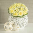 Creamy yellow roses are gathered into a neat dome, their petals brushing one another like a chorus of soft sighs, and are set within a white rattan orb that acts as both bowl and sculpture. Wisps of baby's breath weave through the roses, creating a frothy veil of white that highlights each bloom and lends an airy, garden-fresh lightness to the whole. The arrangement is arranged low and compact, the woven base's crisp lines and open gaps allowing daylight from a nearby window to play across petals and wicker, producing subtle shadows and a luminous edge. Small woven spheres placed to one side introduce a minimalist, almost architectural accent, their rounded forms repeating the bouquet's globe while adding tactile interest. Green blades curl and punctuate the silhouette, providing gentle motion and a hint of organic punctuation. There is a scent of fresh-cut fields and warm linen-a soft, faintly herbaceous undernote beneath the sweet rose perfume-that makes the composition feel lived-in and welcoming in a way familiar to customers around Stanmore tube and The Broadway. As a local florist at Flower Delivery Stanmore, I find this piece is particularly suited to understated celebrations, sympathy gestures, or as an elegant bedside welcome for new parents; it's the sort of arrangement that brings quiet light into a room and reads as both contemporary and comforting.