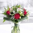 A dramatic red and white bouquet stands proudly in a clear cylindrical glass vase, its stems neatly trimmed and visible through the fresh water, giving an immediate impression of care and freshness. Deep red roses, their velvety petals just beginning to unfurl, share the spotlight with a large, richly ruffled crimson carnation at the front, whose intricate layers create a striking textural focal point. Around them, clusters of pure white and creamy blooms - including white alstroemeria with fine speckles, daisy-like blossoms, and closed white lily buds - soften the intensity of the reds and hint at blossoms still to come. Wisps of feathery baby's breath weave between the larger flowers, like tiny stars scattered through the bouquet, adding volume without heaviness. The arrangement is framed by an abundance of lush greenery: broad, glossy dark green leaves are carefully wrapped and curved around the base like an elegant collar, while slimmer, eucalyptus-style foliage and pointed leaves extend upwards, giving height and gentle movement. The vase is set on a pristine white surface, with a softly blurred, light-filled background that could easily be a bright Stanmore kitchen or living room, catching the gentle daylight that glows across each petal. The scene feels calm yet celebratory, perfect for a romantic anniversary in a home off Stanmore Hill, a birthday surprise near Belmont Circle, or to bring a touch of warmth to an office reception. The bouquet's balanced mix of classic red roses, airy white blooms, and polished greenery creates an atmosphere of thoughtful affection and quietly confident elegance, as if it has just been delivered and carefully placed in pride of position.