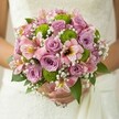 Evoking the serene beauty of Stanmore, this round bridal bouquet bursts with fresh romance and subtle playfulness. Lush lavender-purple and blush pink roses are nestled together, their petals catching gentle light and emitting a soft, sweet fragrance, much like the blooming gardens around Bentley Priory. Splashes of vibrant lime green-button chrysanthemums-bring energy to the arrangement, with each pom-pom adding lively rhythm. Blushing alstroemeria lilies, their petals finely streaked in maroon and gold, accent the bouquet, whispering of faraway places while remaining grounded in Stanmore's heart. Tiny white baby's breath dots the spaces between, their filigree clusters lending an airy magic and a feeling of new beginnings. Glossy deep green leaves provide a natural framework, pulling all the colours into harmony. The bouquet is cradled with care, the bride's hands echoing the gentle spirit of a Stanmore celebration-filled with optimism, tradition, and the quiet elegance of North West London. This arrangement, crafted for lasting memories, makes the perfect companion to joyful promises and the start of a shared future.