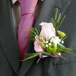 A finely detailed boutonnière is pictured here, resting securely against the deep tone of a formal suit jacket, the kind worn for weddings, engagements, or formal dinners in Stanmore and the surrounding areas. At its heart is a single rose of soft, pale pink, just beginning to open, with inner petals curled delicately toward the centre in an elegant spiral. The rose is embraced by an arrangement of fresh greenery, each leaf placed to create a layered, textural effect: some are narrow and linear, others slightly curved, giving the composition a sense of balance and flow. Interwoven between the leaves are small sprays of tiny white gypsophila flowers, their delicate clusters appearing almost cloud-like, adding a feeling of lightness and romance. Just below the rose, a green button chrysanthemum provides a crisp, contemporary twist, its rounded form and vivid lime shade drawing the eye and contrasting beautifully with the softer tones above. The stems at the base of the boutonnière are neatly gathered and expertly wrapped in a subtle lavender-mauve ribbon, which extends slightly below in a small, satin tail that softens the overall line. The arrangement looks freshly made, the rose petals smooth and unblemished, the foliage a rich, healthy green as though it has just been prepared for same-day delivery around Stanmore, Kenton, or Edgware. You can imagine a florist from Flower Delivery Stanmore pinning this piece to a lapel with quiet pride, knowing it will be worn close to the heart during vows, speeches, and shared laughter. The overall mood conveyed is one of intimacy, refinement, and gentle celebration, perfectly suited to milestone moments.