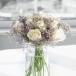 A full, rounded bouquet of creamy white roses and lilac statice stands at the centre of the frame, its elegance heightened by soft natural light that feels like a calm morning in Stanmore. Around a dozen white roses, each bloom perfectly formed and just beginning to open, reveal layers of velvety petals with a gentle ivory warmth at their hearts. Between the roses, airy clouds of tiny lavender-purple statice flowers thread delicately in and out, their papery texture and fine stems adding a feathery, almost mist-like halo that softens the composition. The flowers are gathered in a tall, clear cylindrical glass vase, the stems neatly aligned and visible through the water, a vibrant green lattice that hints at their freshness. The vase rests on a bright white surface, likely a table by a window in a Stanmore home, catching and reflecting the daylight so that faint, soft-edged shadows fall around the base. The background is blurred and pale, suggesting an uncluttered, peaceful interior where this bouquet might be the quiet focal point of the room. The overall effect is serene and uplifting: a design that would feel at home in a tranquil living room overlooking Stanmore Country Park, or on a bedside table bringing comfort after a difficult day. The roses suggest a gentle, clean fragrance, with a whisper of green from the stems and statice, evoking a spring garden after rain. This arrangement feels well suited to thoughtful sympathy gestures, understated anniversary surprises, or simply to bring a touch of refined calm to everyday life in the local neighbourhood.