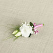 On a neutral, textured background that could easily be a linen cloth in a Stanmore florist's workroom, a refined boutonniere is placed with care, waiting to be pinned to a lapel. The central focus is a large white lisianthus flower, beautifully unfurled, its petals generously ruffled and softly curved, reminiscent of a delicate piece of fabric gently turned by hand. The creamy-white tone of the bloom gives it an inviting warmth, avoiding any harsh brightness. Tucked lovingly at its side are two lisianthus buds, slender and elongated, still closed in anticipation, embodying the idea of love just beginning to unfold. Sprigs of baby's breath are threaded among the blooms, their tiny white blossoms forming a light, cloud-like frame that adds an airy, almost dreamlike quality. Fine strands of green foliage and reedy stems arc around the flowers, providing contrast and a touch of garden freshness, as if gathered from greenery near Bentley Priory Nature Reserve. The gathered stems at the base of the boutonniere are tightly wrapped in a vivid pink satin ribbon, the wrap smooth and even, giving a polished, finished look. The satin has a gentle sheen that catches the light when viewed from different angles, bringing a modern, joyful splash of colour to the composition. A small, carefully tied bow completes the design, its neat loops showing the steady hand of an experienced florist at Flower Delivery Stanmore. Together, the soft petals, delicate buds, airy baby's breath, and bright ribbon create a boutonniere that feels romantic, fresh, and perfectly suited to weddings, proms, and special celebrations across Stanmore and the nearby hills.