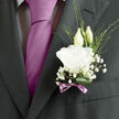 In this close-up image, the groom's lapel becomes a backdrop for a small yet expressive wedding boutonnière, crafted with the kind of quiet attention you might expect from a florist in Stanmore preparing flowers for a ceremony near Stanmore Country Park or along Marsh Lane. The focal point is a single white lisianthus bloom, its petals opening in layered, ruffled waves that feel both soft and assured, like finely pressed fabric touched by gentle afternoon light. Just behind the open flower, two slender lisianthus buds reach upwards, their pale green and ivory forms still tightly closed, symbolising the unfolding journey about to begin. Surrounding the lisianthus, petite clusters of baby's breath create a delicate, cloud-like aura, their countless tiny blossoms gleaming like dewdrops scattered around the central bloom. Wispy strands of fresh green foliage thread between the flowers, lending a sense of natural movement, while a single polished leaf is shaped and tucked beneath, deep green and glossy, giving the boutonnière a grounded, architectural feel. The stems are bound with a fine mauve ribbon, tied into a neat bow that introduces a hint of lavender colour, gently echoing the groom's tie without overpowering it. There is an imagined whisper of scent-clean, faintly sweet, and fresh-drifting from the lisianthus and baby's breath as the groom moves through his wedding day. The piece feels calm, confident, and carefully considered, a small floral accent that marks the importance of the moment while reflecting the relaxed, welcoming character of Stanmore celebrations among close family and friends.