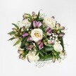 A top-down view captures a compact, hand-tied bouquet laid against a spotless white background, the kind of clean, bright setting you might find on a polished kitchen table in a Stanmore home. At the heart of the arrangement, creamy white and soft ivory roses unfold in various stages of bloom, from tight buds with a whisper of buttery yellow at their centres to fully opened flowers whose velvety petals spiral outwards in generous layers. Threaded between the roses are elegant alstroemeria, their narrow petals washed in mauve, lavender, and berry-pink tones, some still enclosed in smooth green buds, others half-open to reveal fine, painterly streaks of deeper colour and touches of fresh green. Delicate sprays of white gypsophila, like tiny clouds of froth, drift between the larger blooms, softening every transition and giving the whole bouquet an airy, romantic lightness. Around the base, broad, glossy leaves in a deep, woodland green cradle the flowers, their folded lines adding structure and contrast, much like the calm, leafy streets that frame Stanmore's parks and gardens. The lighting is even and gentle, with no harsh shadows, highlighting the freshness of each petal and the crispness of the foliage. The bouquet feels poised between classic elegance and contemporary simplicity, well-suited to birthdays, quiet anniversaries, or thoughtful get-well wishes. It evokes the feeling of a cool spring morning walk past local gardens, and suggests the careful eye and practiced hands of an experienced florist who understands how to balance colour, texture, and emotion in a single, harmonious design.